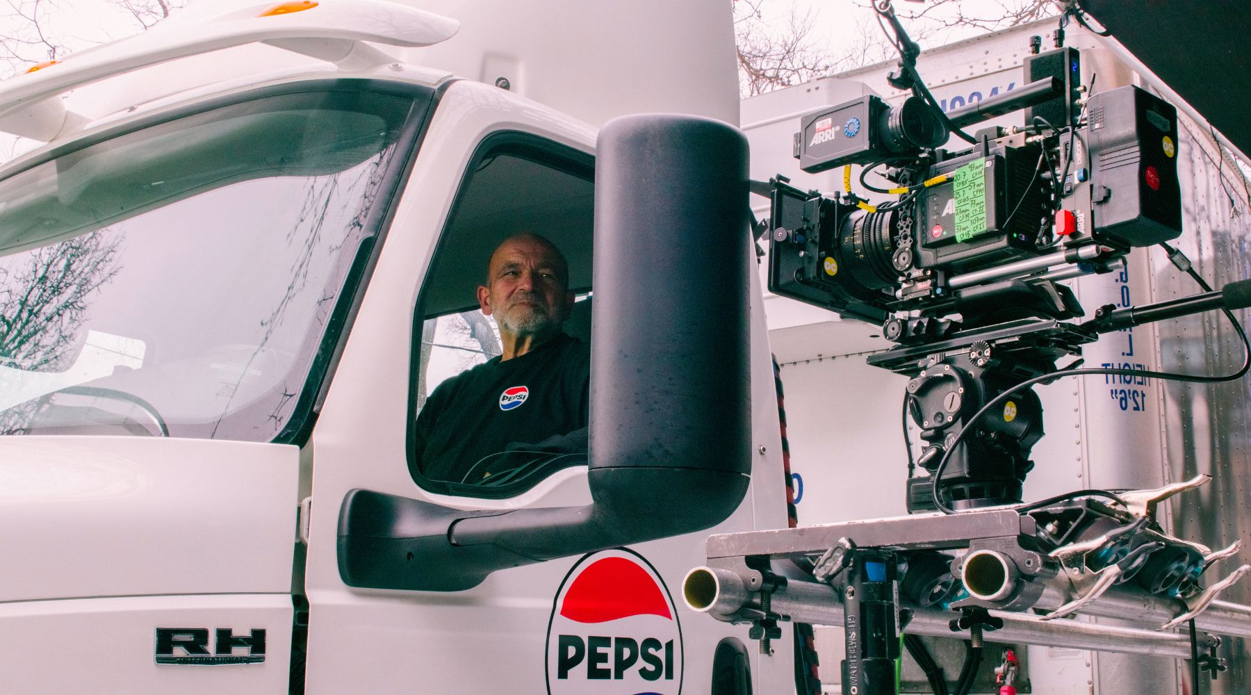  Un chauffeur de PepsiCo Canada est assis dans la cabine d’un camion blanc portant le logo Pepsi, tandis qu’une caméra professionnelle fixe filme la scène à traver la fenêtre ouverte côté conducteur pendant le tournage d’une publicité. 