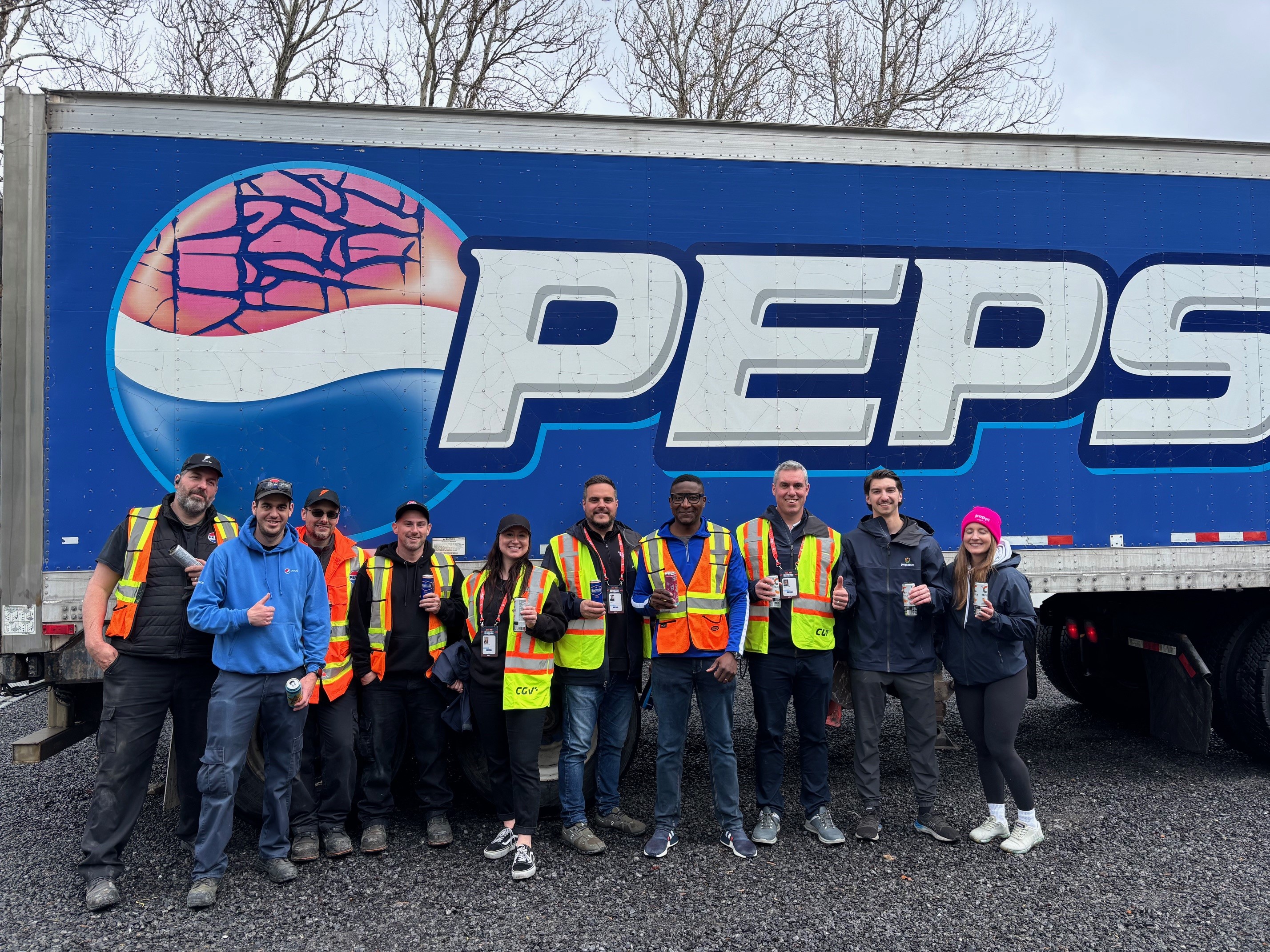 PepsiCo team members in safety vests stand together holding beverages in front of a large Pepsi-branded transport truck outdoors.