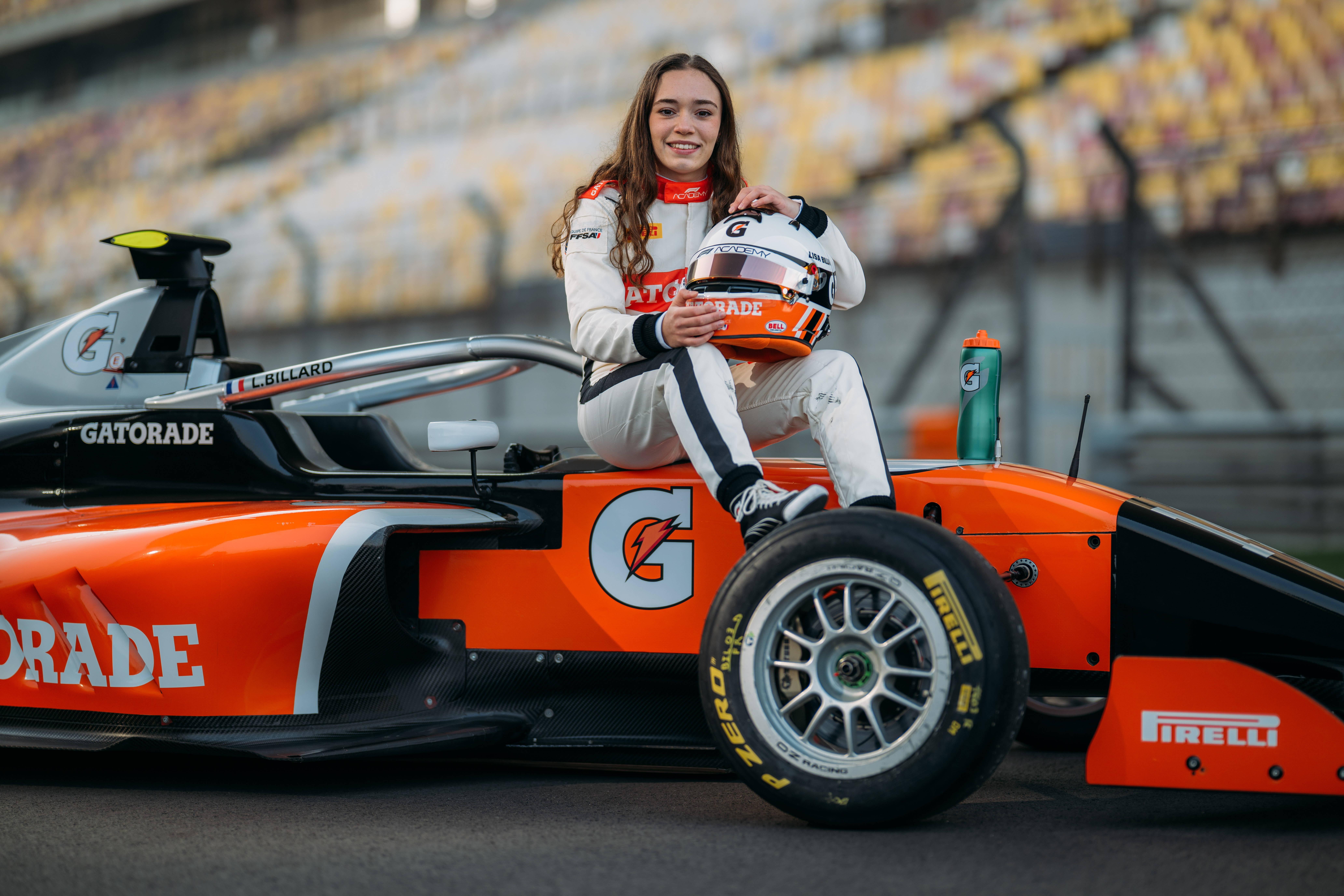 Race car driver sits on an orange Gatorade-branded formula car, holding a helmet on a racetrack with stadium seating in the background.